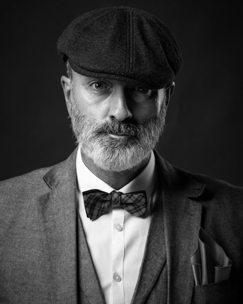 A bearded man wearing a flat cap, grey jacket, white shirt and tartan bow tie against a dark background.