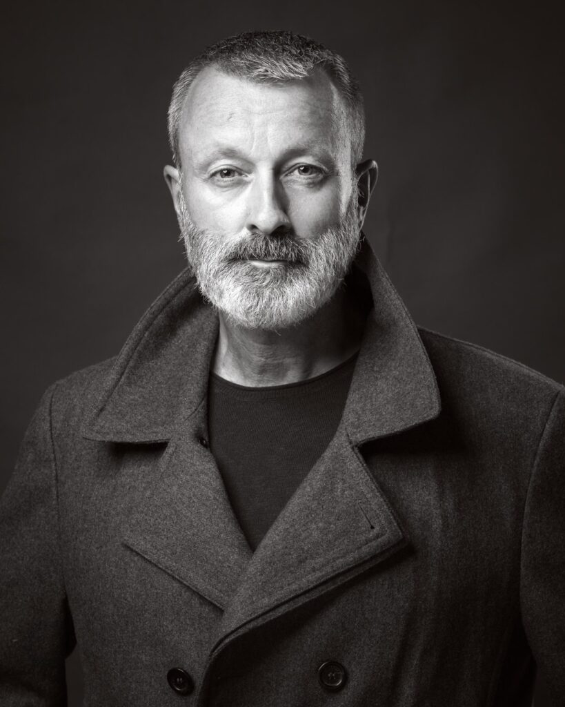 A man with grey hair and beard wearing a dark wool coat over a black shirt, photographed against a dark background.