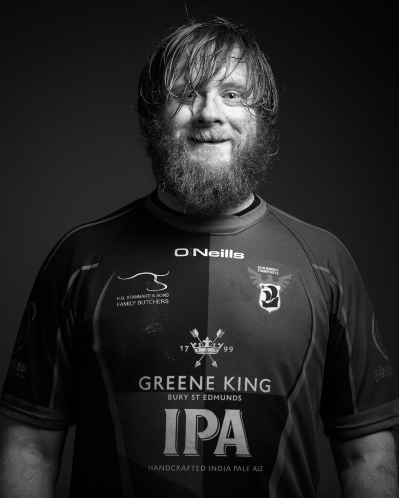 A bearded man with shoulder-length hair wears a black O'Neills sports shirt featuring Greene King IPA branding against a dark background.