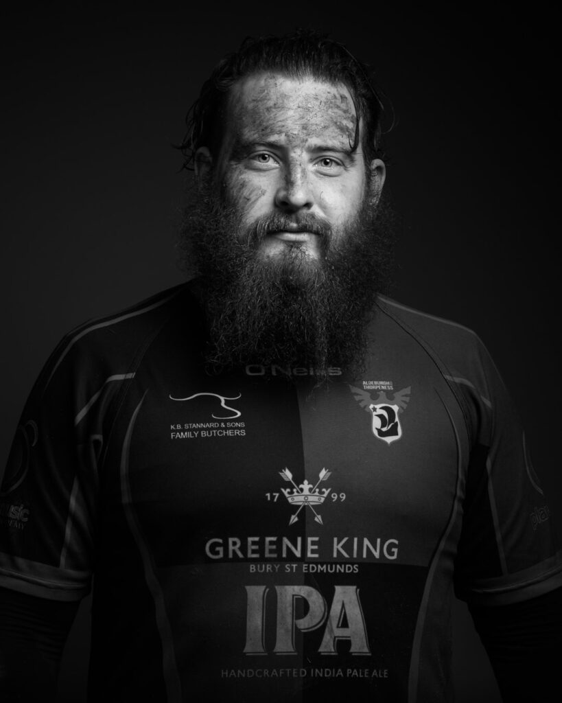 A bearded man in a dark sports jersey stands against a black background in a portrait photograph.