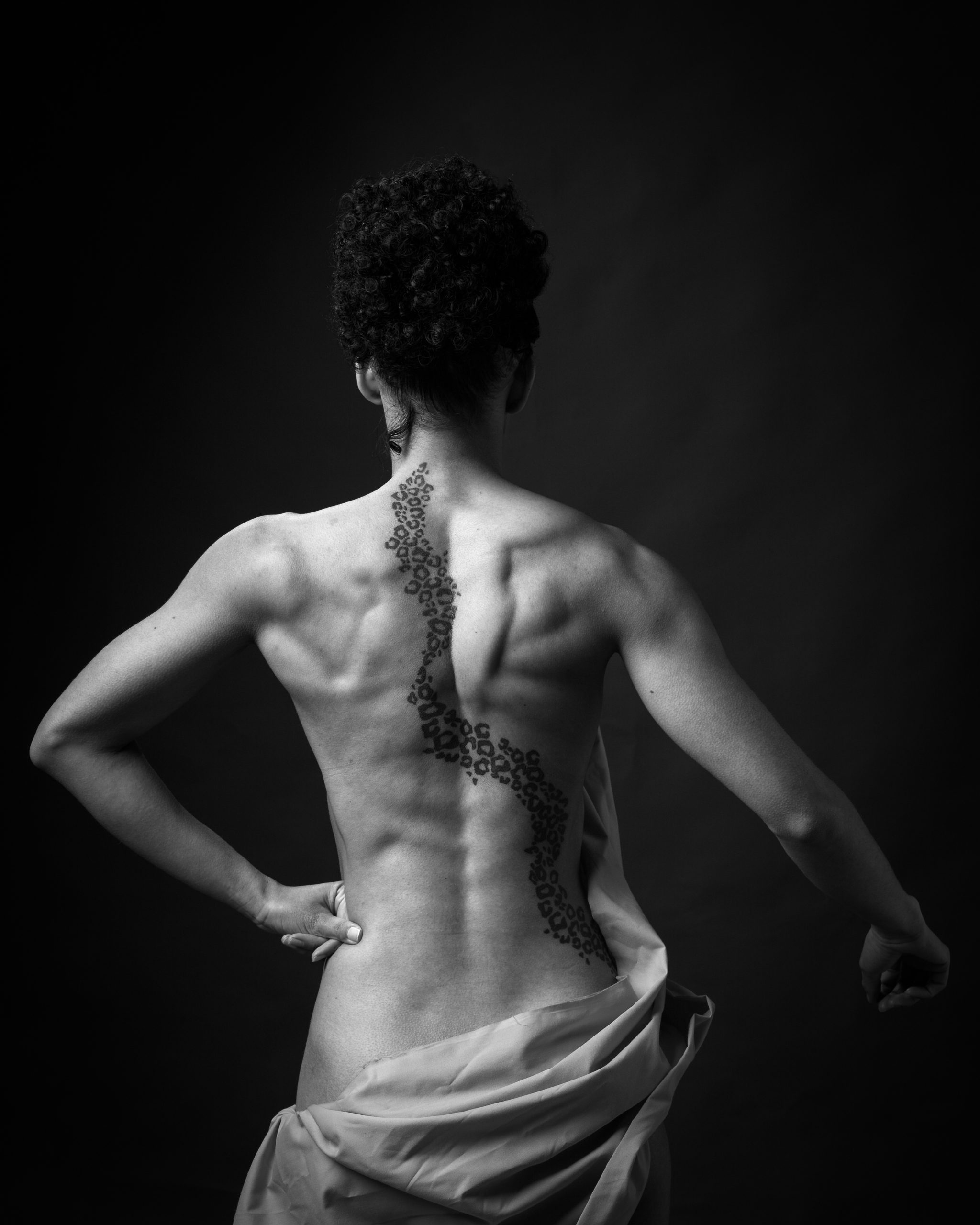 A person photographed from behind against a black background, displaying a decorative tattoo running down their spine.