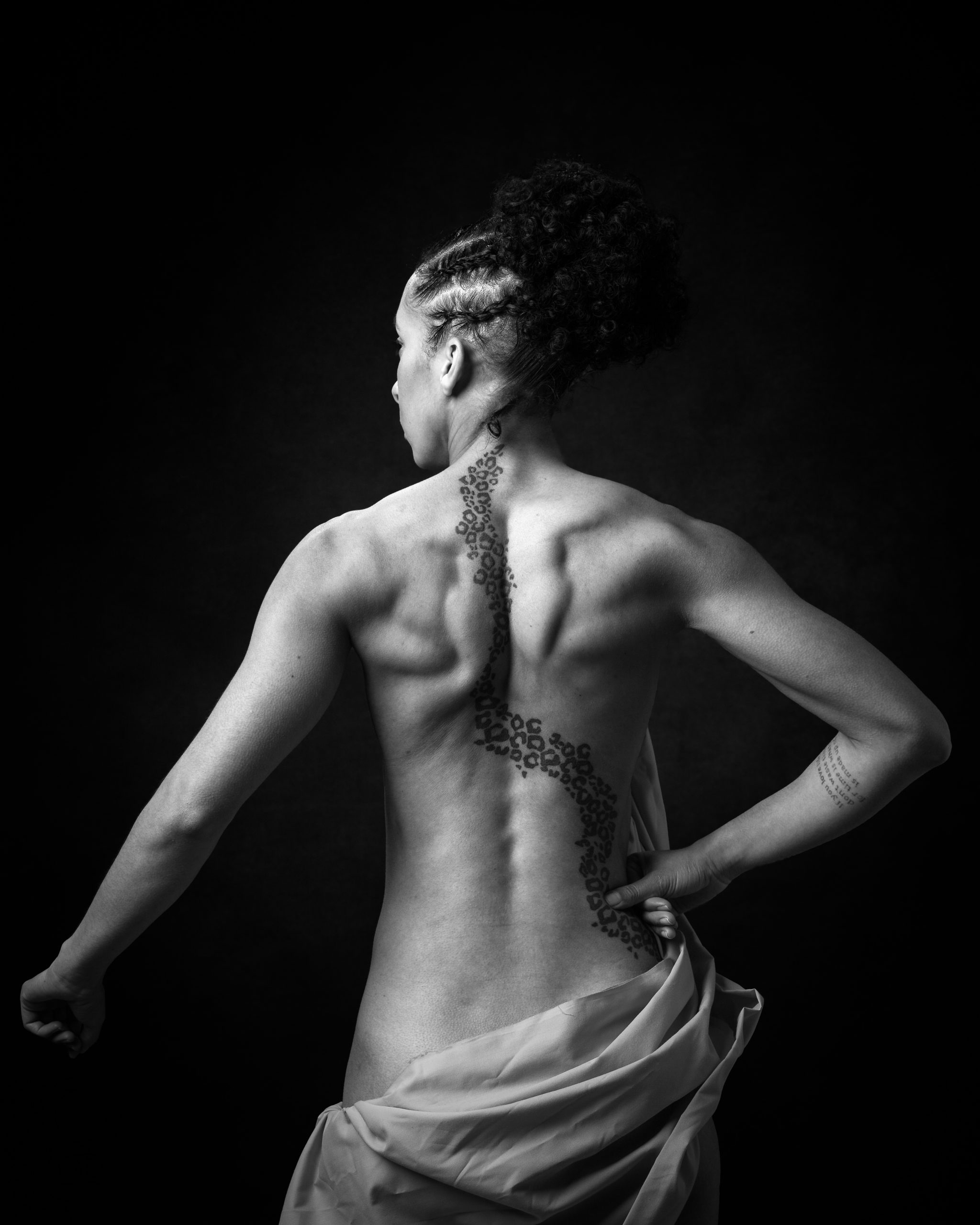 A person shown from behind against a black background, displaying back tattoos and wearing a draped light fabric around their waist.