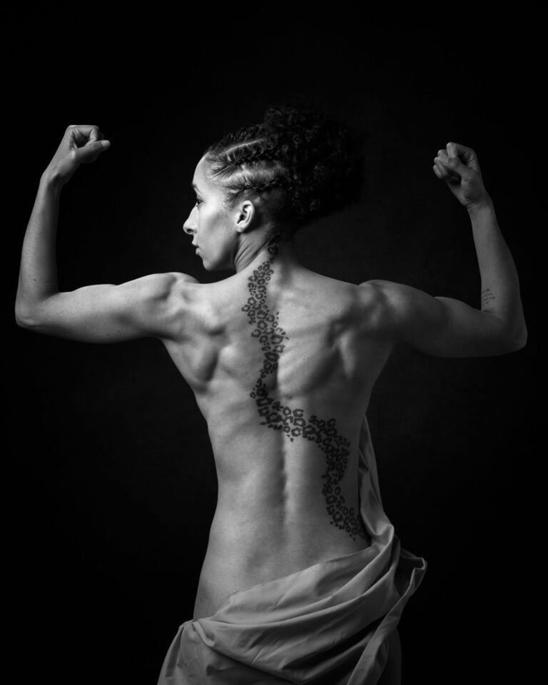 A person shown from behind flexing their arms, displaying a decorative tattoo down their spine, against a black background.