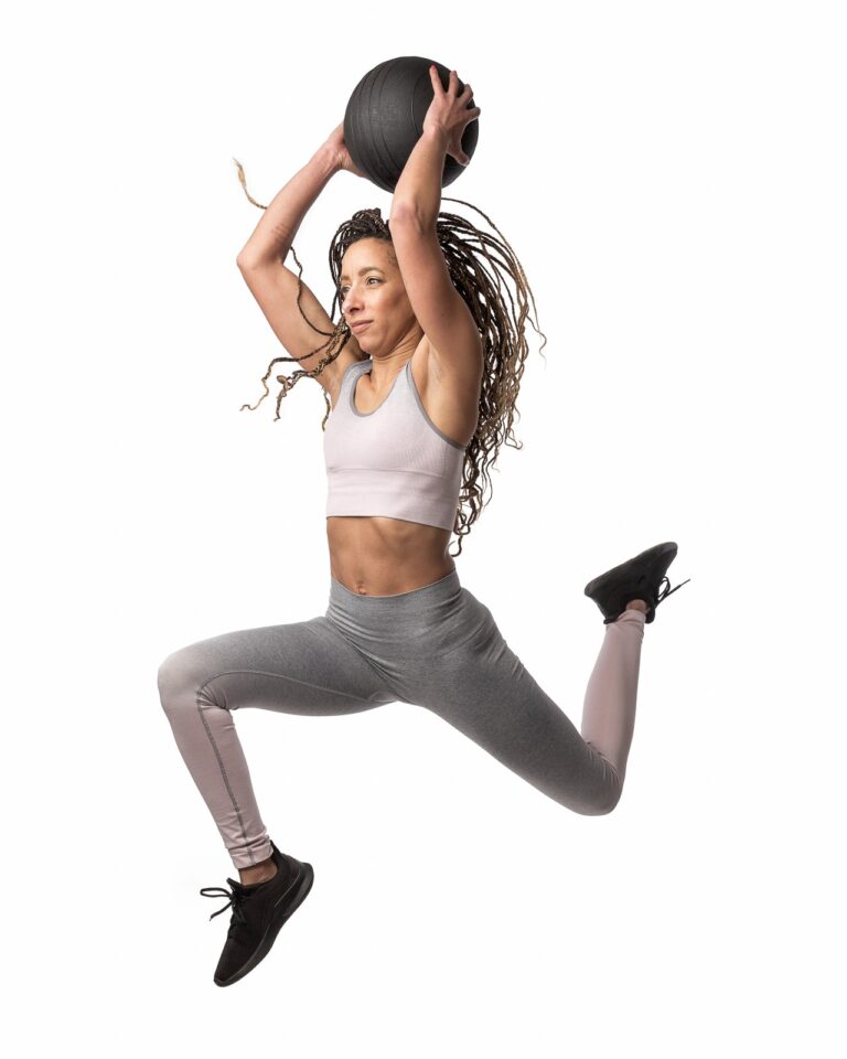 A woman in athletic wear performs a medicine ball slam exercise while jumping against a white background.