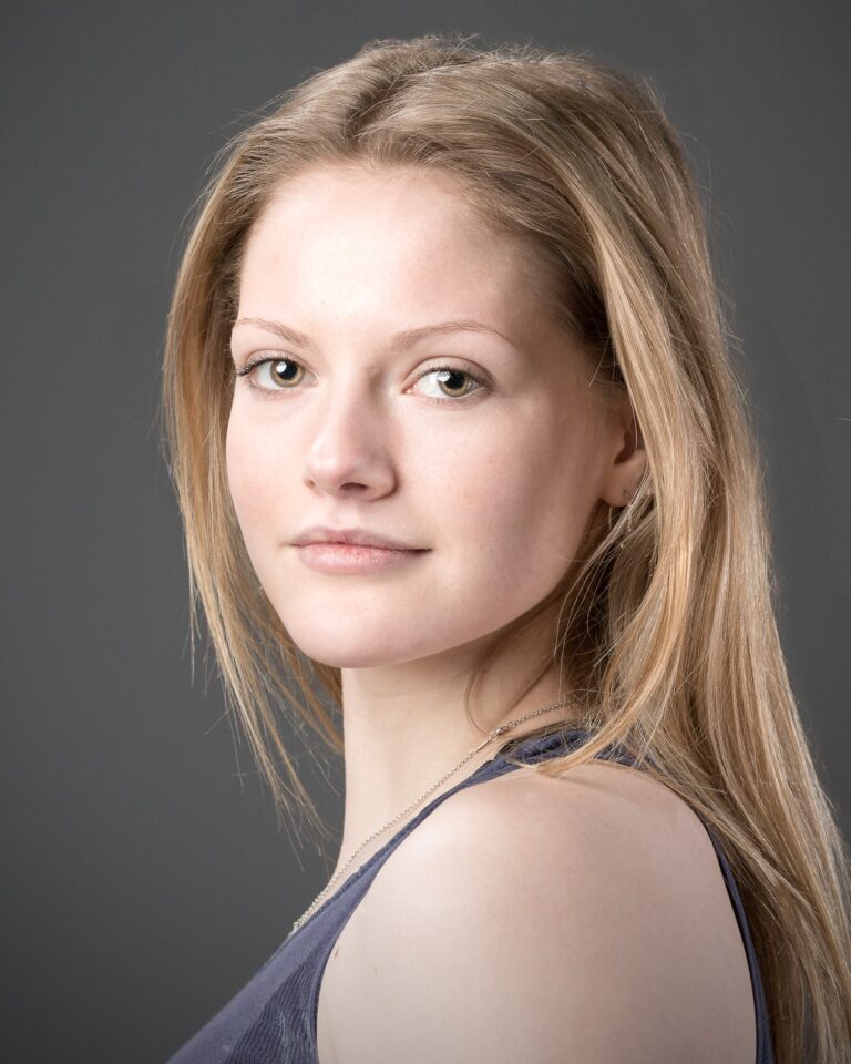 A woman with long, straight brown hair looks towards the camera against a plain grey background. She wears a dark blue top.