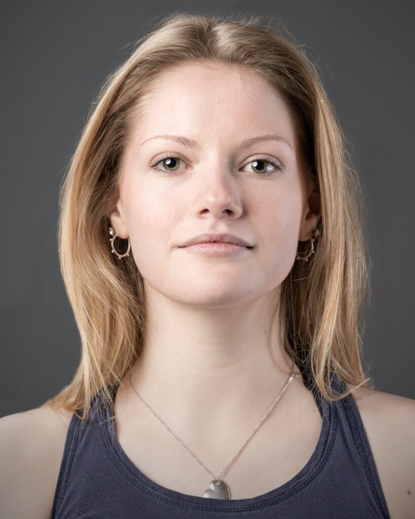 A woman with shoulder-length auburn hair wearing a dark tank top and pendant necklace looks directly at the camera against a grey background.