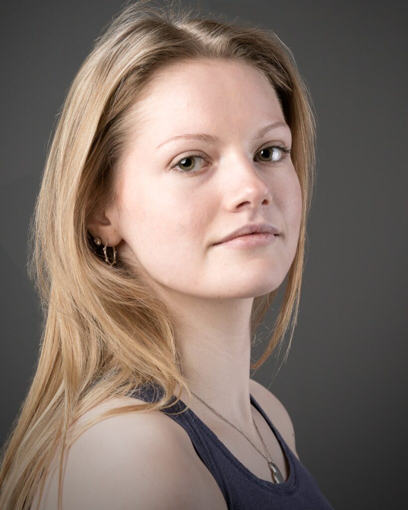 Portrait of a woman with light brown hair, wearing a dark top and necklace, looking over her shoulder against a grey background.