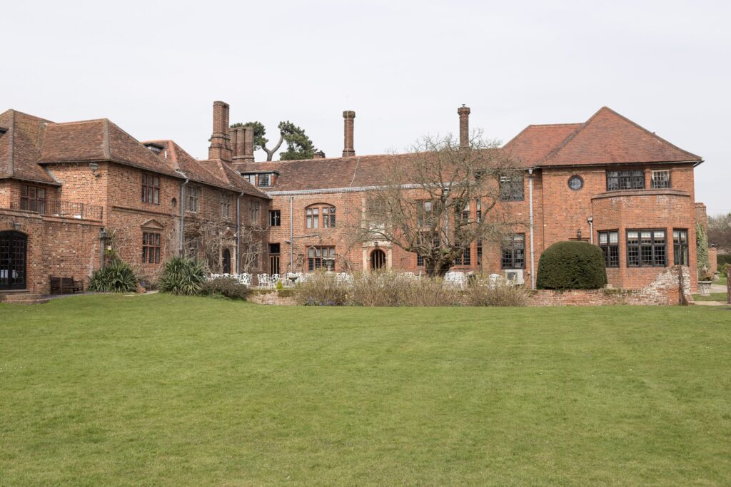 A large red brick mansion viewed from across a manicured lawn, with multiple chimneys and gabled roofs visible against an overcast sky.