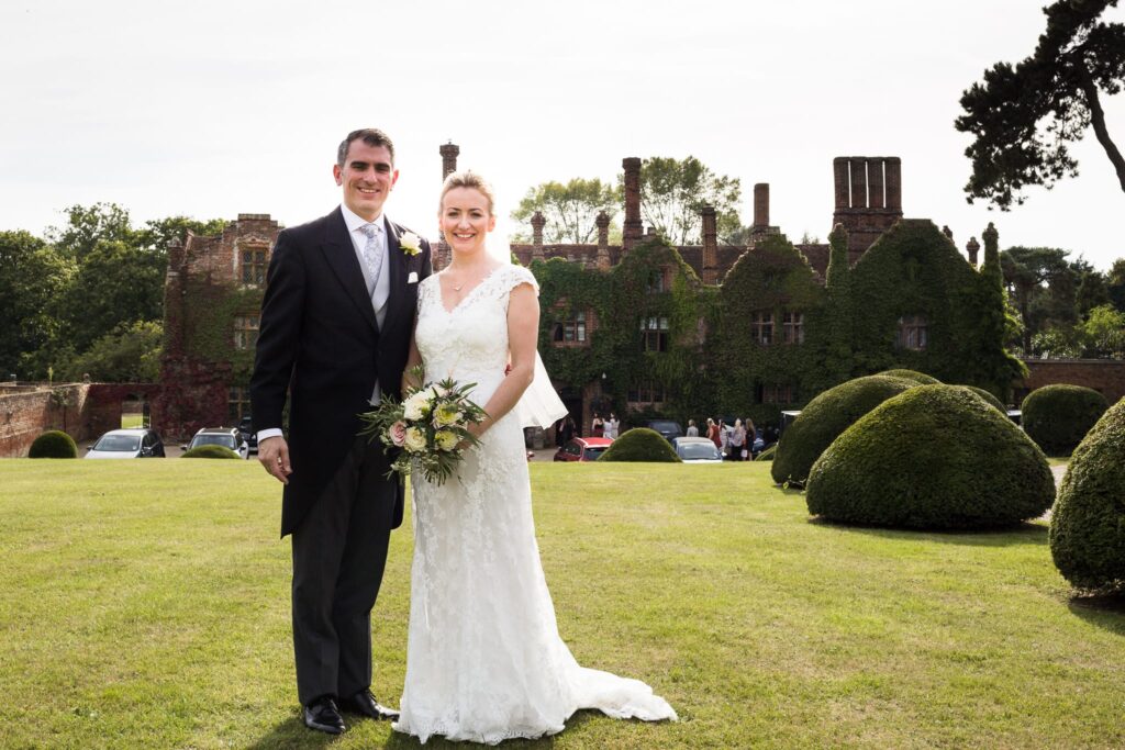 A bride and groom stand together on a manicured lawn in front of a large ivy-covered castle, smiling at the camera.