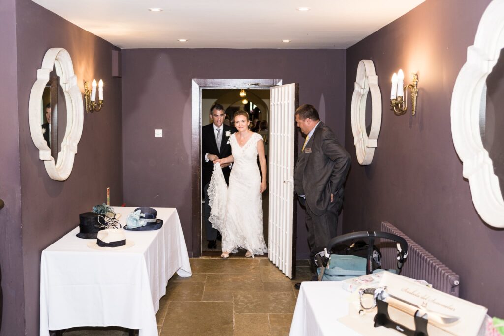 A bride and groom enter a purple-walled room with a man in a suit greeting them. Various items are on tables nearby.