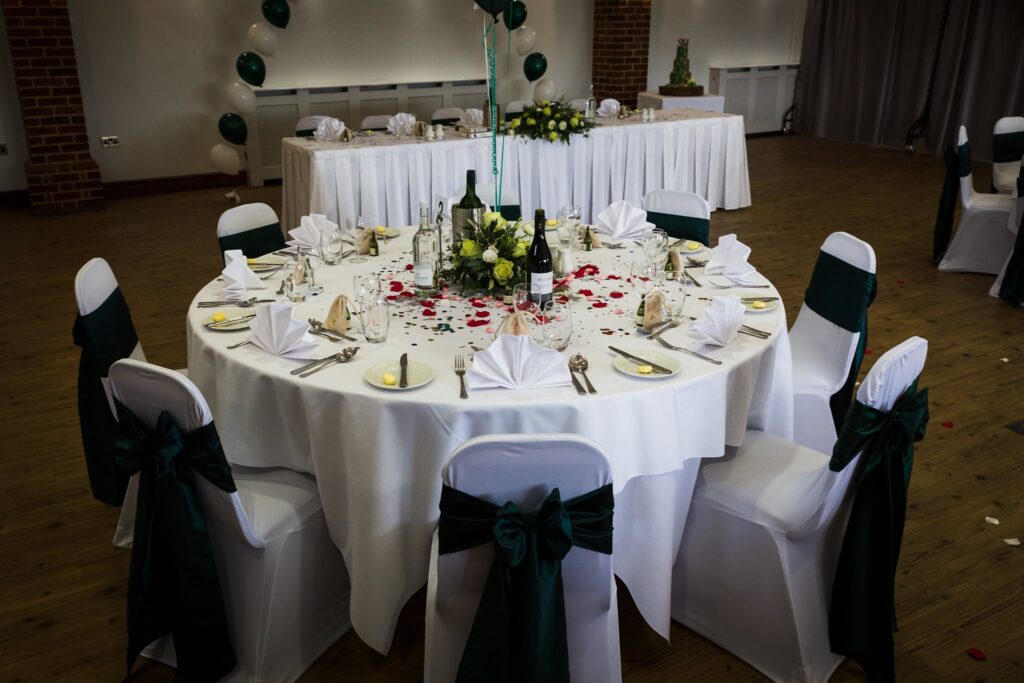 A formal wedding reception setup with a round table decorated in white, dark green, and gold with place settings and floral centrepiece.