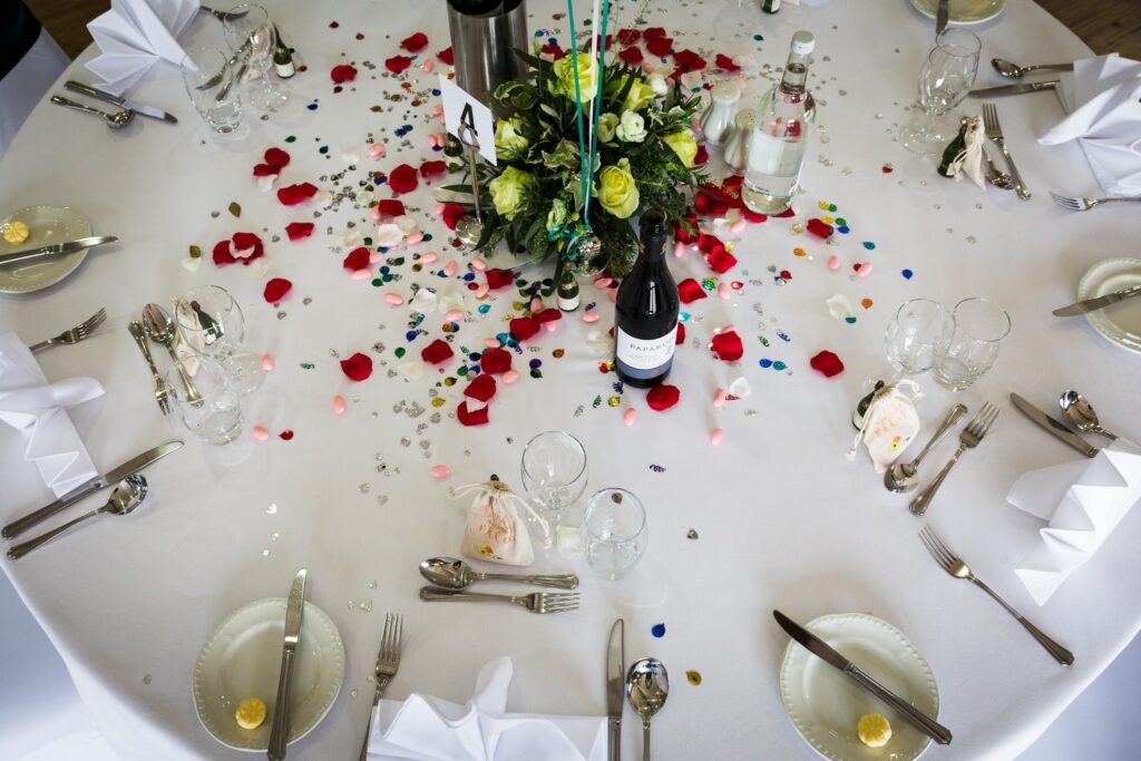 A round banquet table set for a formal dinner with white tablecloth, place settings, and a floral centrepiece surrounded by rose petals and confetti.