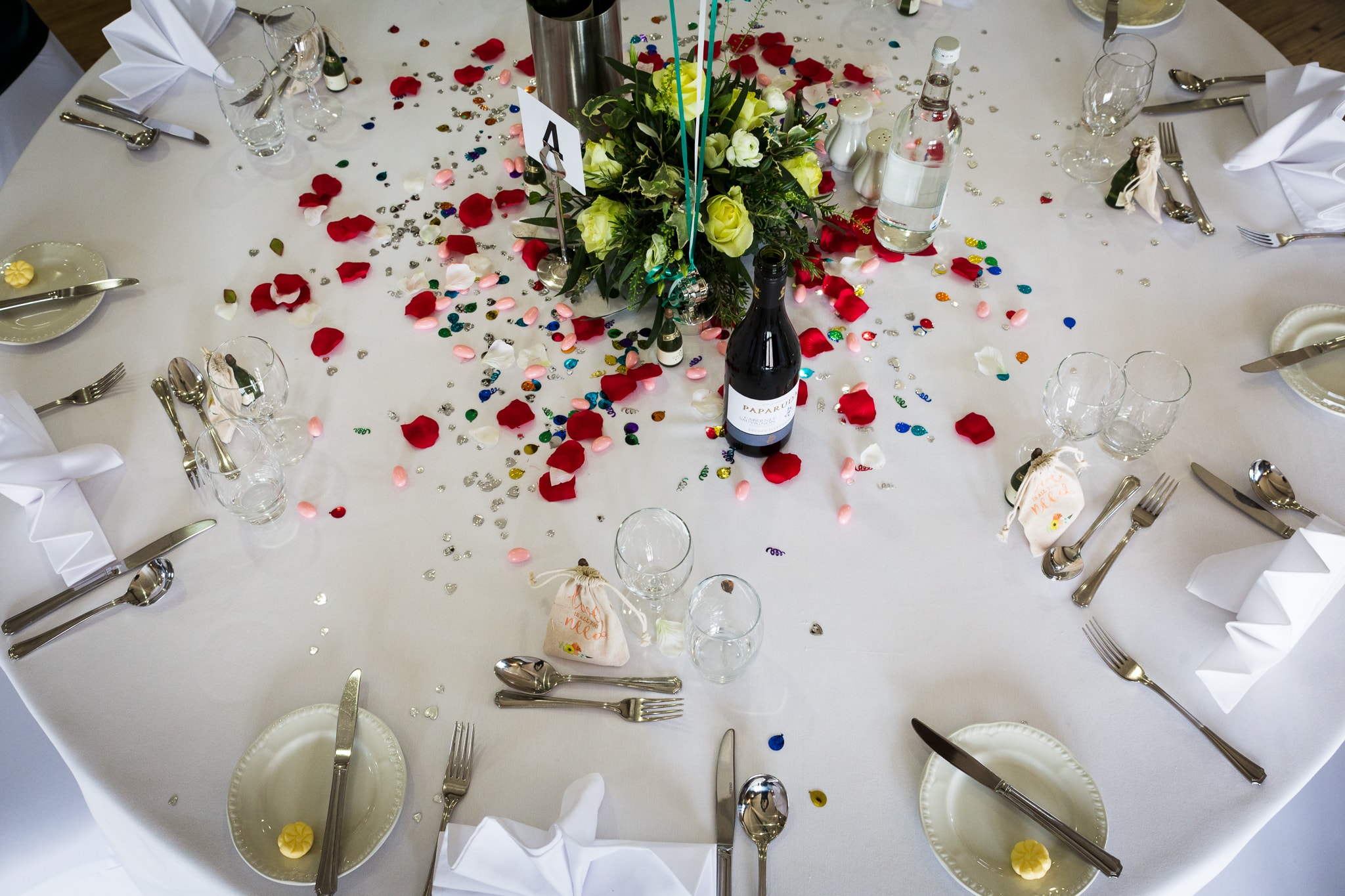 A round banquet table set for a formal dinner with white tablecloth, place settings, and a floral centrepiece surrounded by rose petals and confetti.