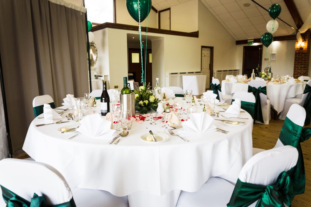 An elegantly decorated banquet hall with round tables dressed in white linens, green chair sashes, floral centrepieces, and green balloons suspended from the ceiling.