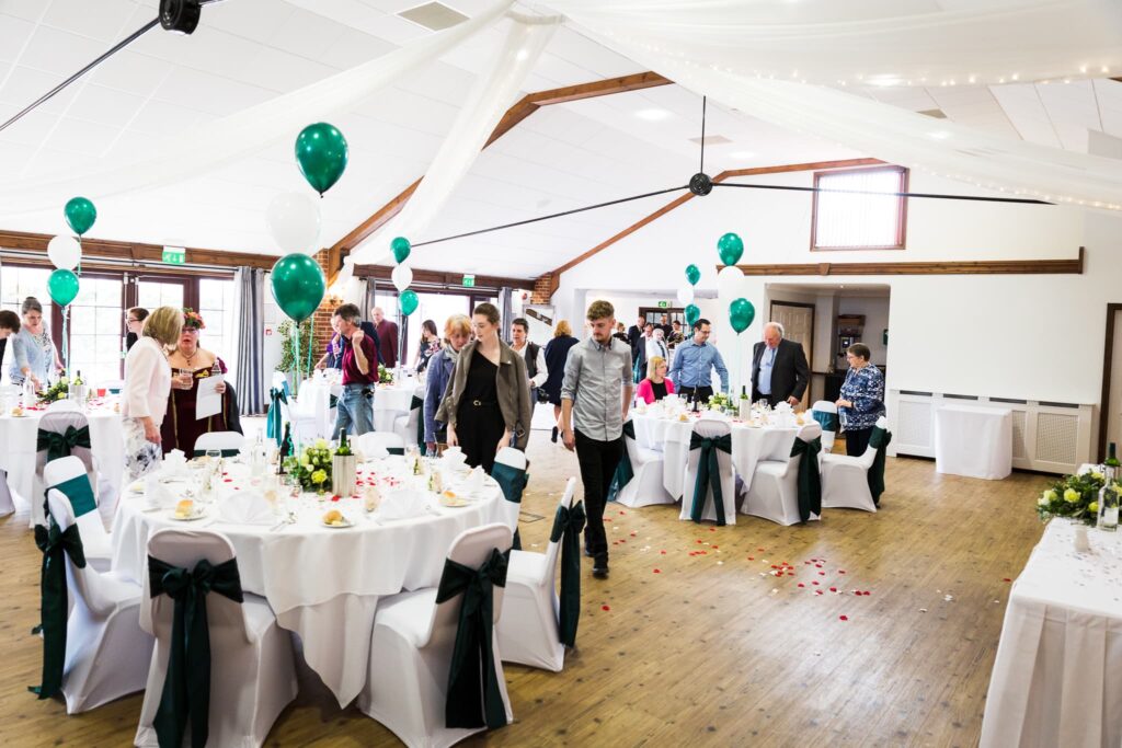 A wedding reception in a hall with round tables, green and white balloons, and guests mingling among the decorations.