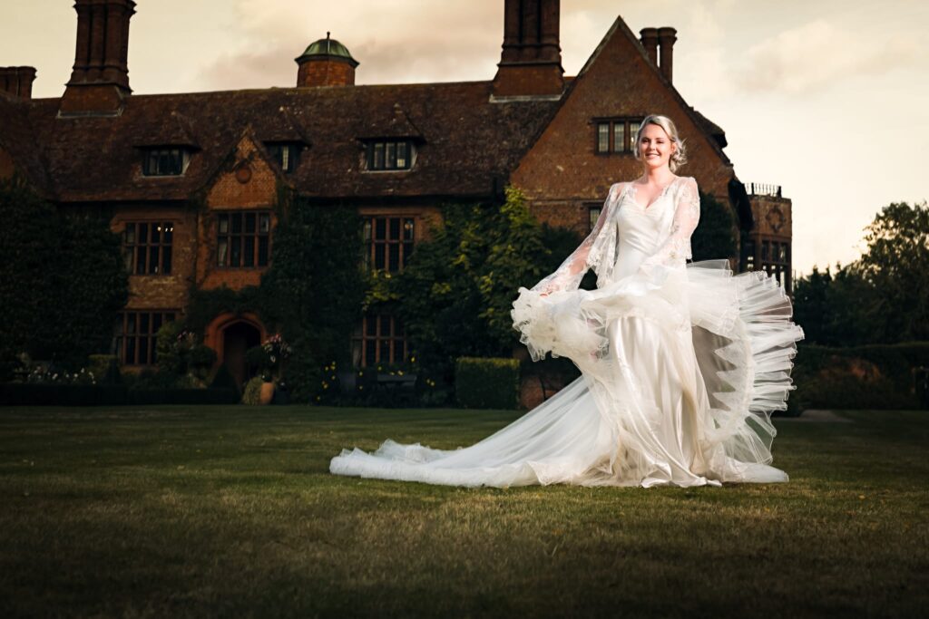 A bride in a flowing white wedding dress with a long train stands on a lawn before a grand brick manor house.
