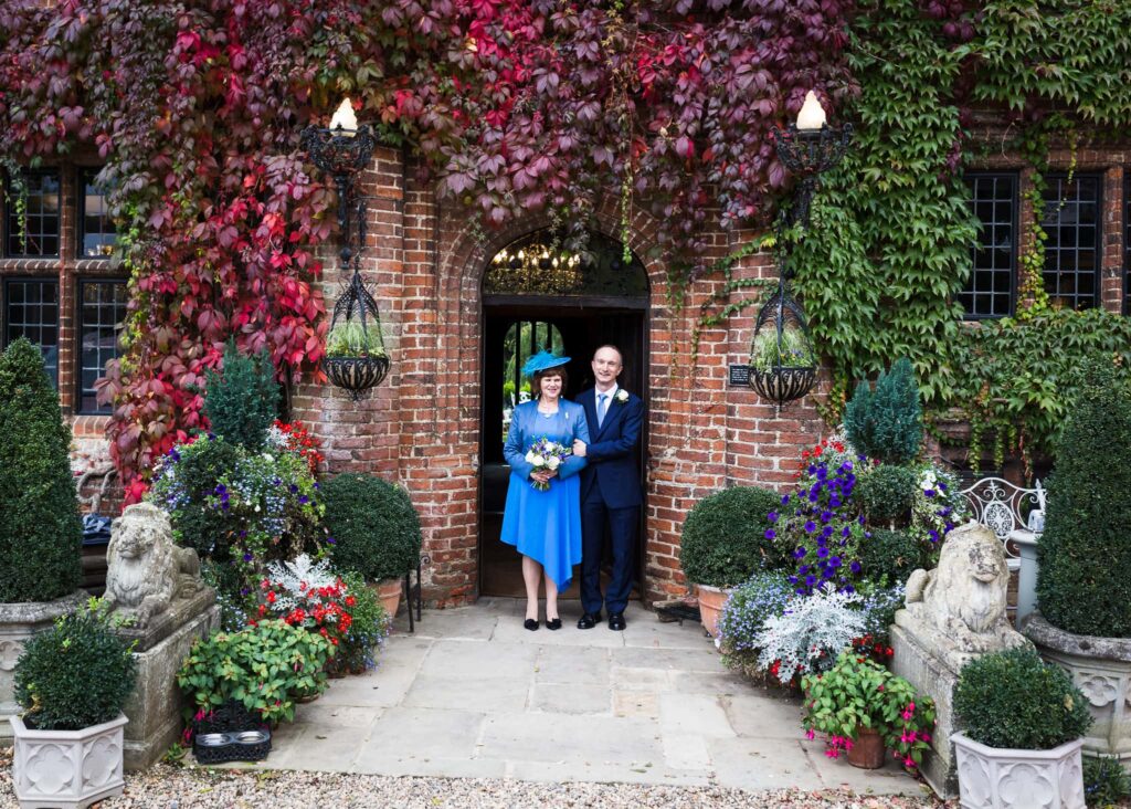 A woman in a blue outfit and hat stands with a man in a dark suit at a brick archway entrance, surrounded by vibrant red and green climbing ivy.