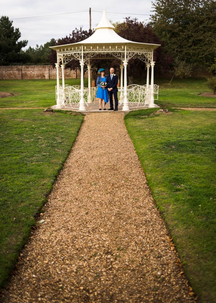 A woman in a blue dress and a man in a dark suit stand together inside an ornate white gazebo in a manicured garden.