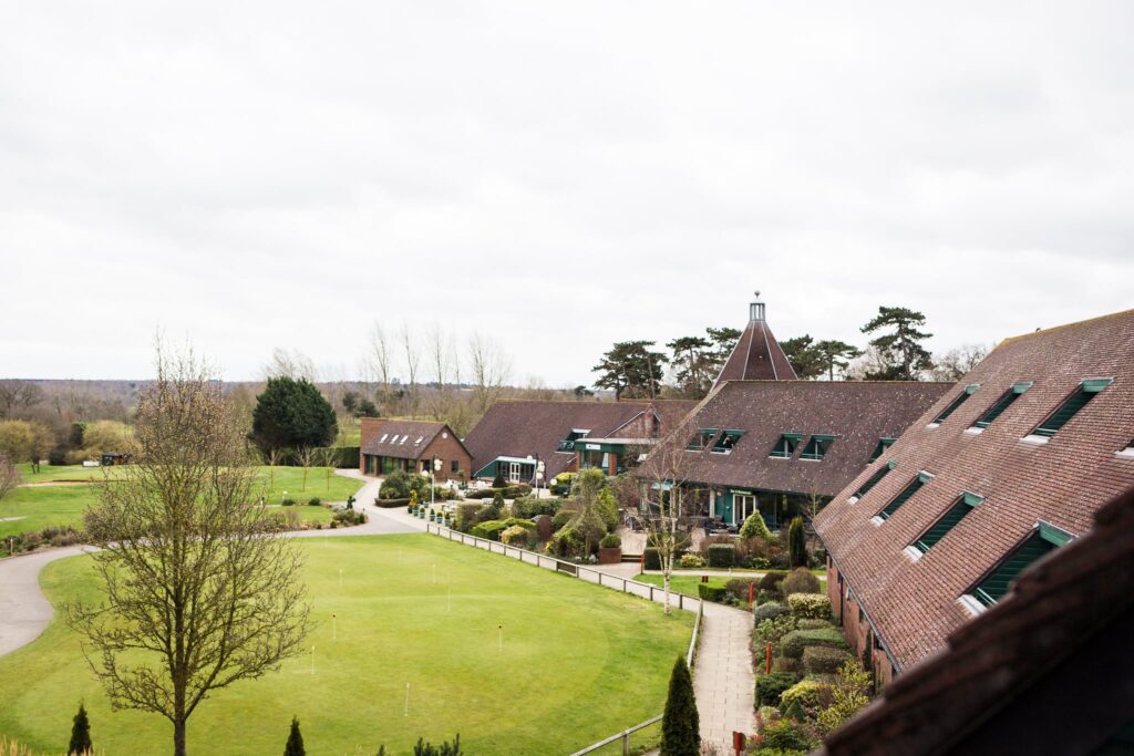 A sprawling countryside estate with brick buildings, manicured lawns, and a distinctive spire, surrounded by green fields under an overcast sky.