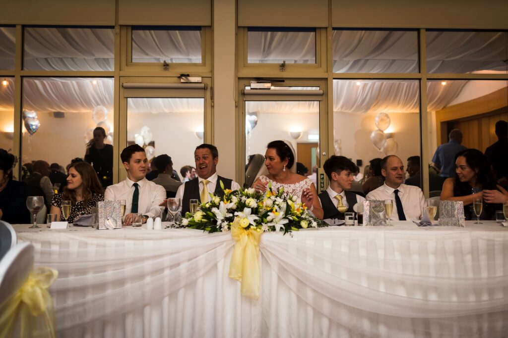 Wedding reception with guests seated at a long table decorated with white draping and yellow and white floral centerpiece.