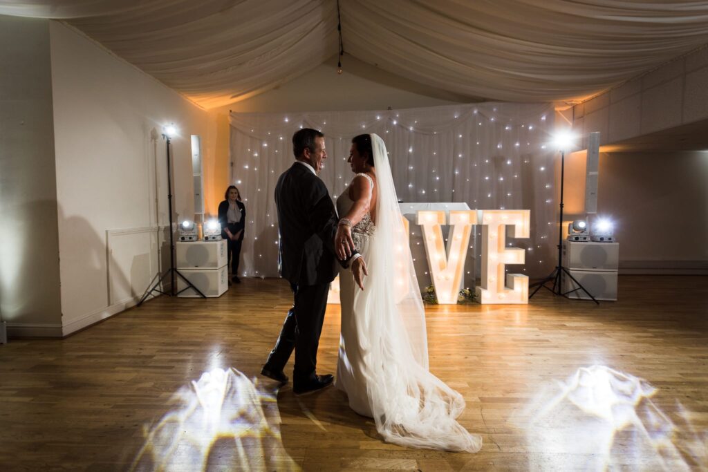 A bride and groom dance together in an elegant venue with fairy lights and a large illuminated LOVE sign behind them.