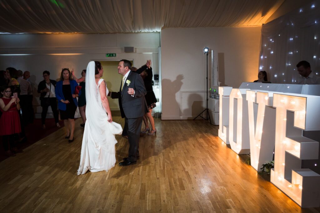 A bride and groom dance together at their wedding reception in a decorated venue with warm lighting and illuminated letters nearby.