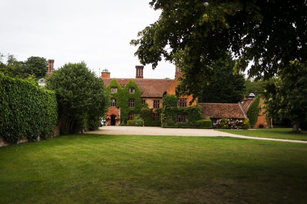 A brick manor house covered in ivy, viewed across a lawn with mature trees framing the scene on either side.