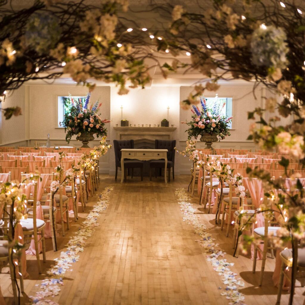 A decorated church aisle for a wedding ceremony, lined with chairs, flowers, and petals leading to the altar.