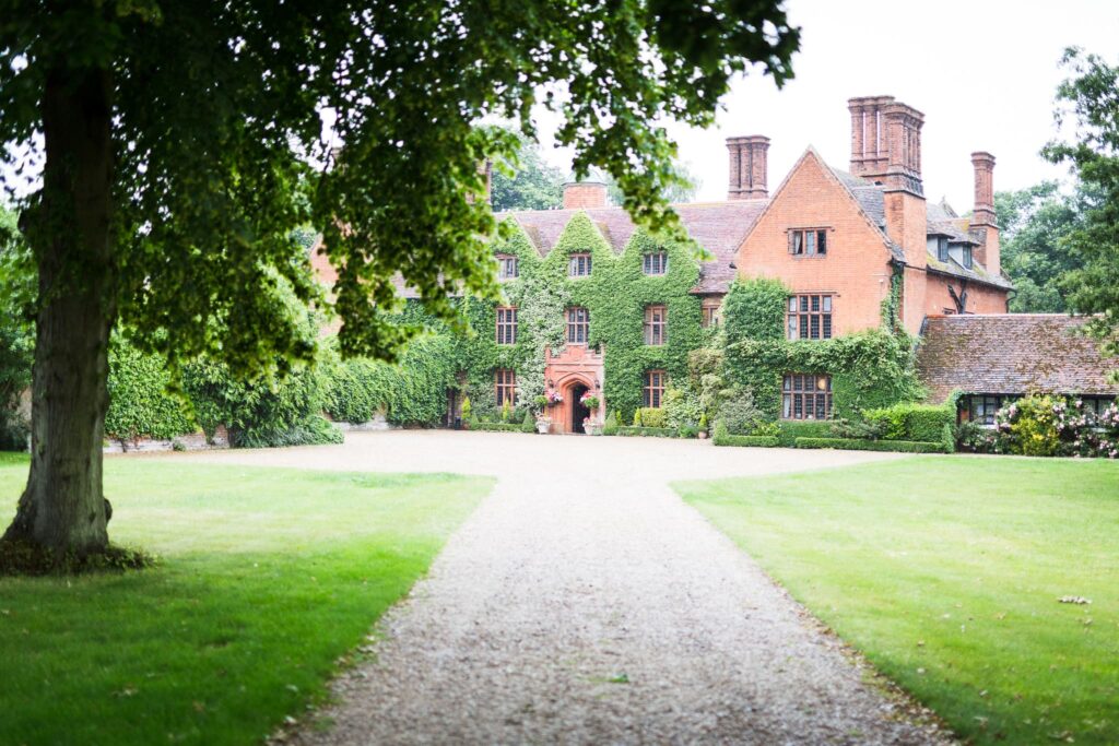 A gravel driveway leads toward a large red-brick country house covered in ivy, flanked by mature trees and manicured lawns.