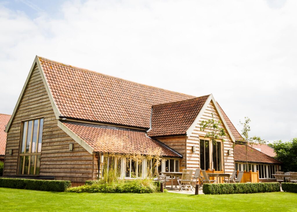 A contemporary timber-clad house with terracotta tile roofs and large windows, featuring a patio with wooden furniture and manicured lawn.