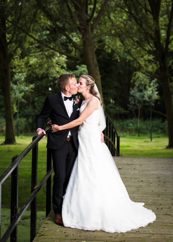 A bride and groom kiss on a wooden bridge in a tree-lined park. The groom wears a black tuxedo and the bride wears a white lace wedding dress.