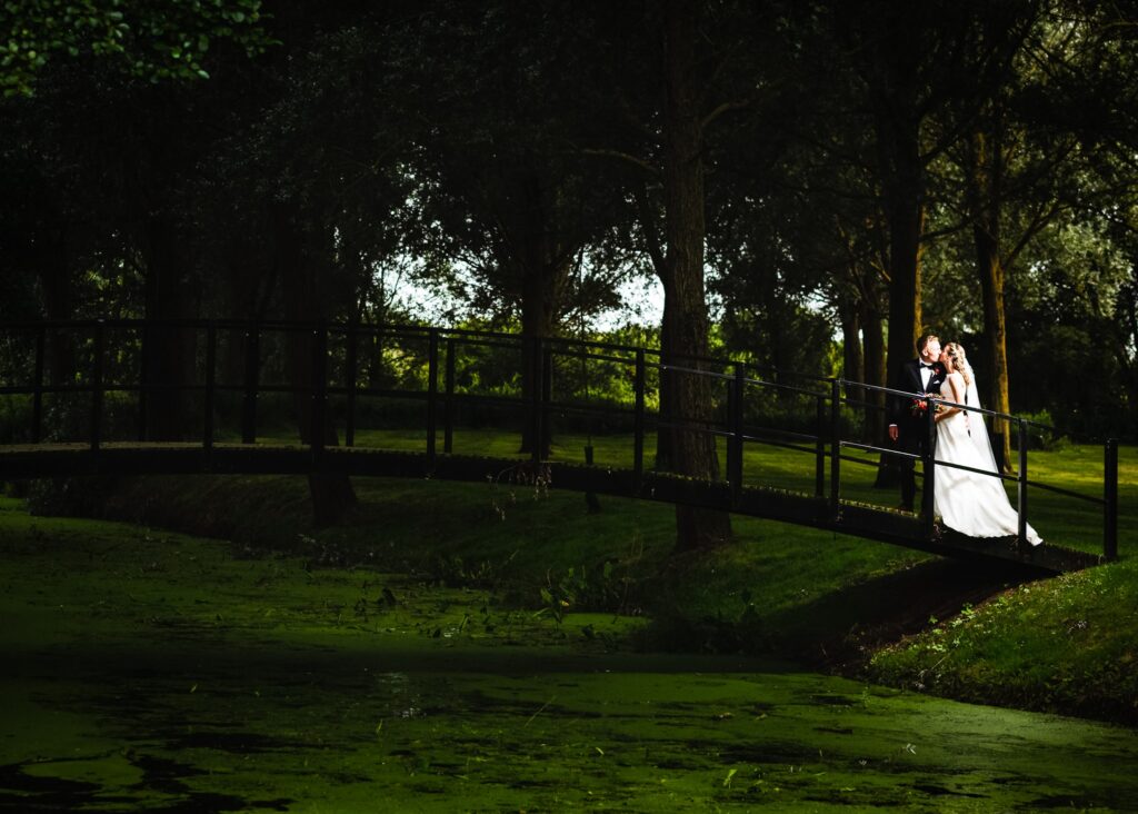 A bride and groom stand together on a wooden bridge over a pond, surrounded by tall trees in a lush green park setting.