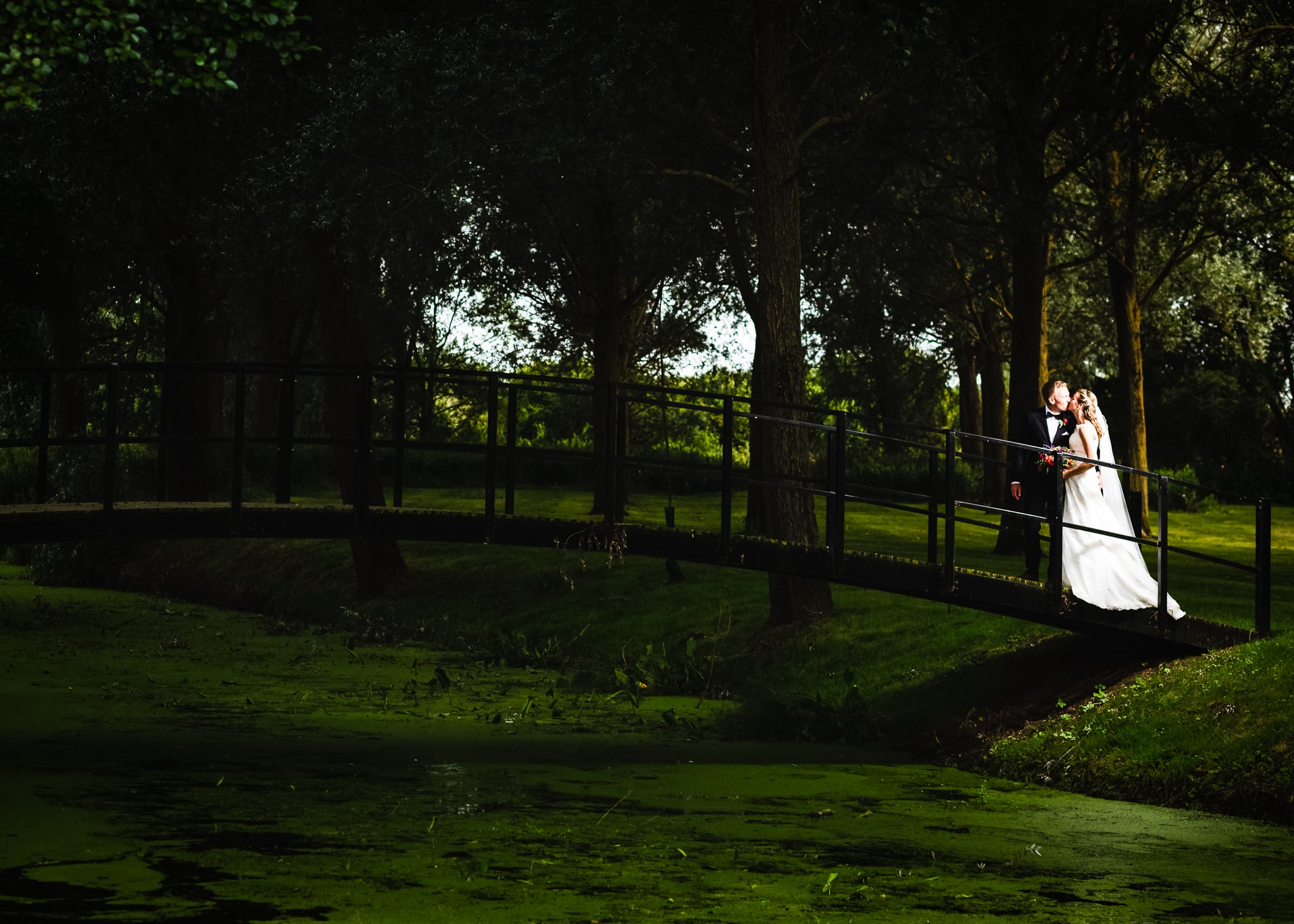 A bride and groom stand together on a wooden bridge over a pond, surrounded by tall trees in a lush green park setting.