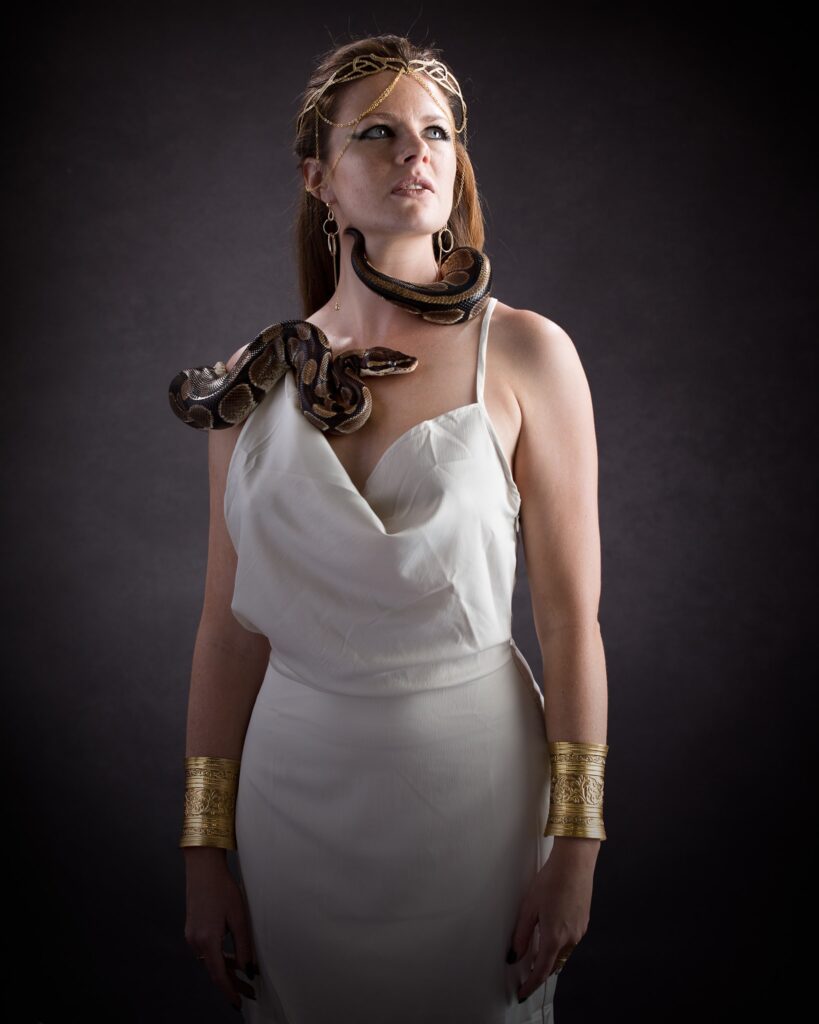 Woman in white dress with snakes around neck and shoulder against dark background, wearing gold accessories.
