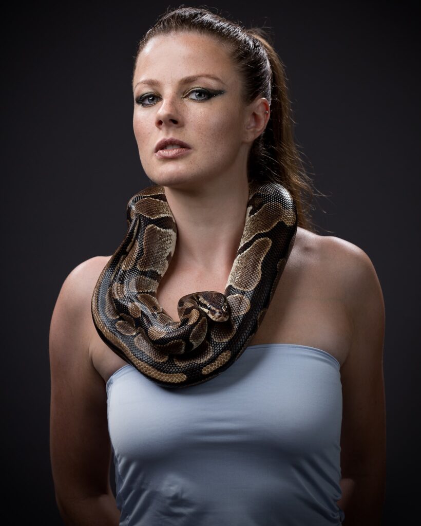 Woman wearing light blue top with a ball python draped around her neck and shoulders against a black background.