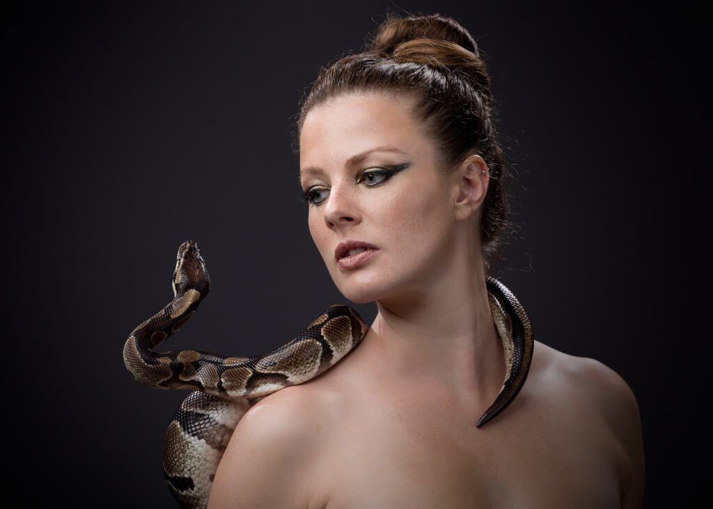 Woman with hair in a bun, looking downward, with a large snake draped across her shoulder against a dark background.