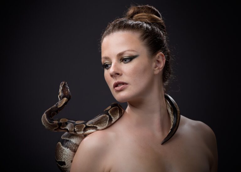 Woman with hair in a bun, looking downward, with a large snake draped across her shoulder against a dark background.