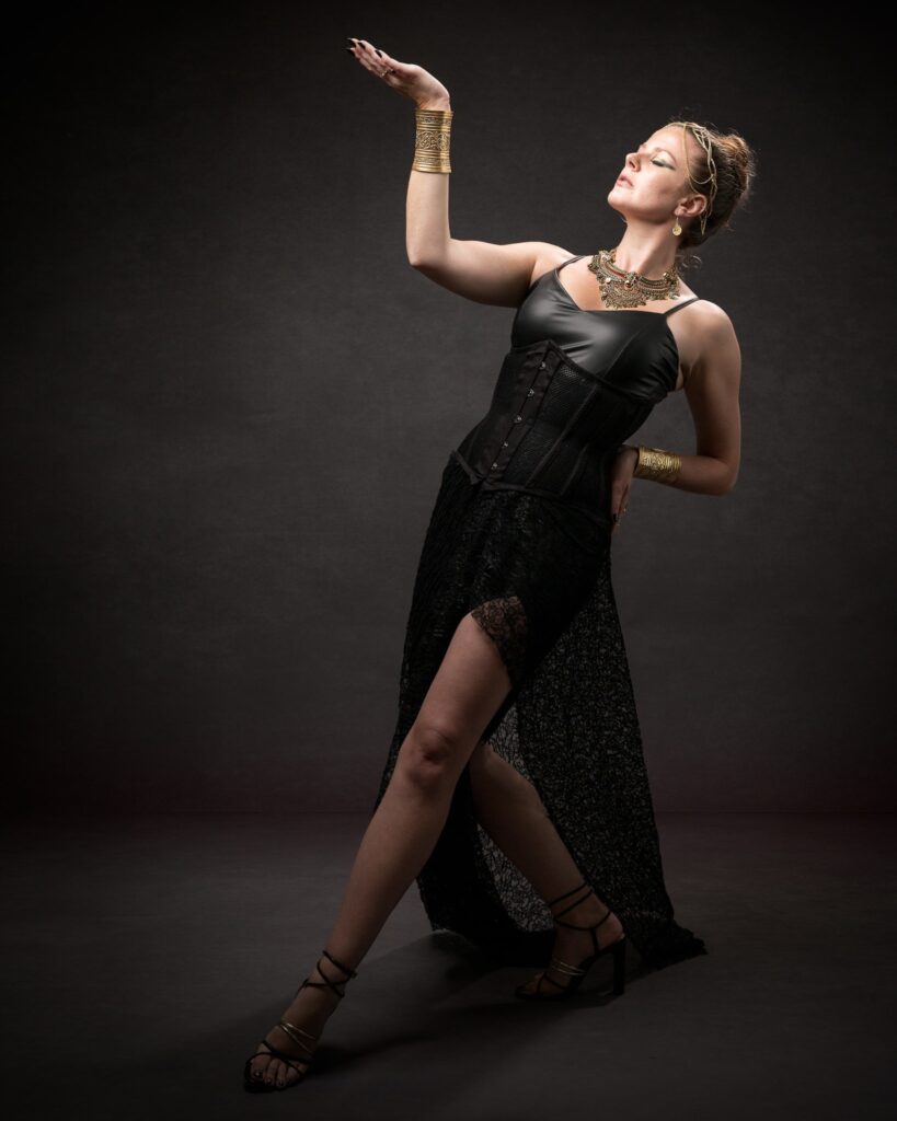 A woman in a black corset dress and gold accessories poses against a dark background with her arm raised.