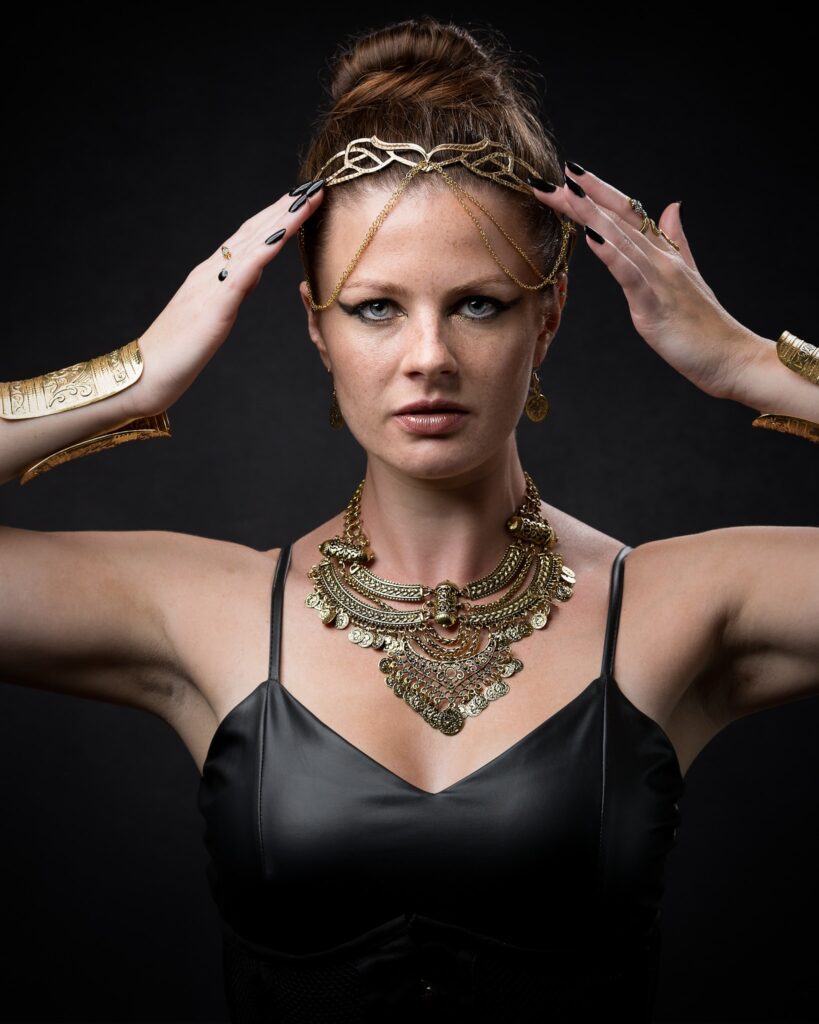 A woman with dark hair in a bun wears elaborate gold jewelry and a black top against a dark background.
