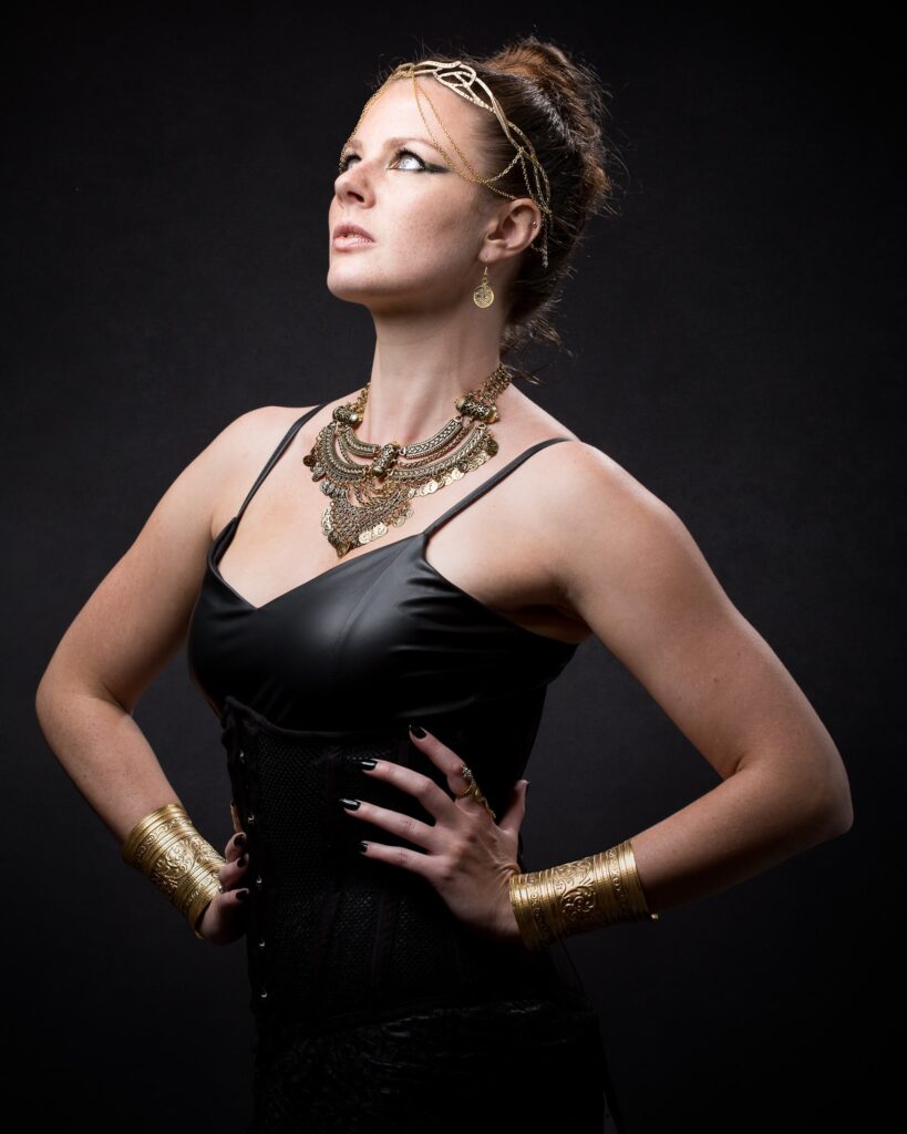 Woman in black dress with gold jewelry against dark background, looking upward in profile pose.
