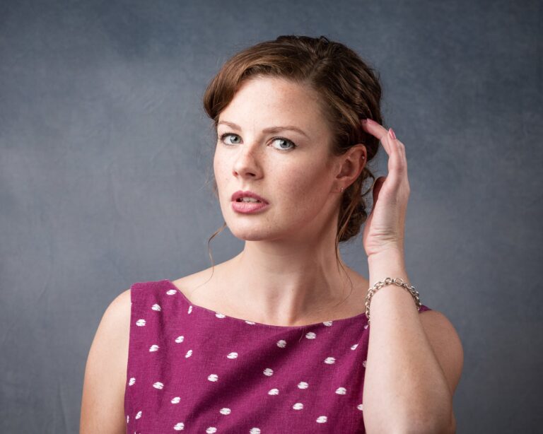 A woman with brown hair styled in an updo wearing a burgundy top with white polka dots, hand raised to her ear, against a grey background.