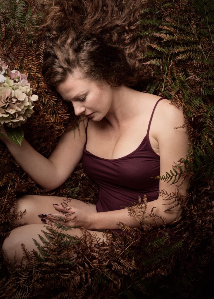 A woman in a burgundy top reclines among ferns and flowers, eyes closed in a peaceful pose.