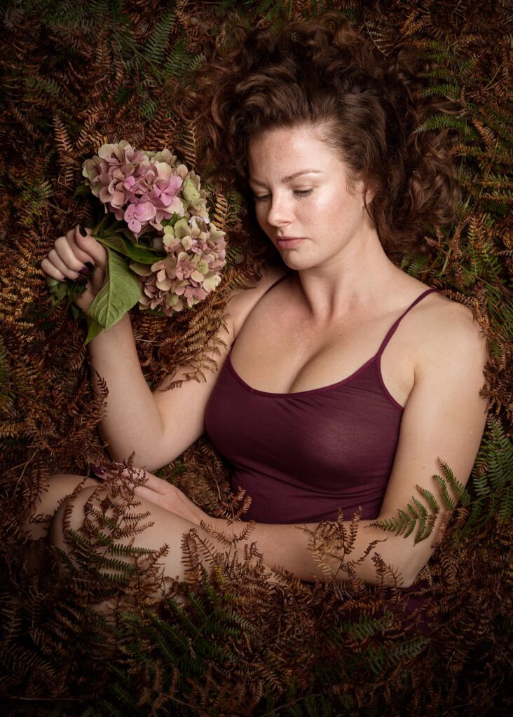 Woman in burgundy top holding pink hydrangeas, surrounded by ferns and dried foliage, gazing downward.