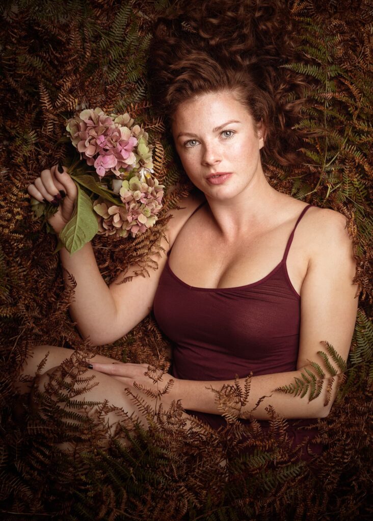 Woman holding pink hydrangea flowers, wearing burgundy top, surrounded by dried ferns and foliage backdrop.