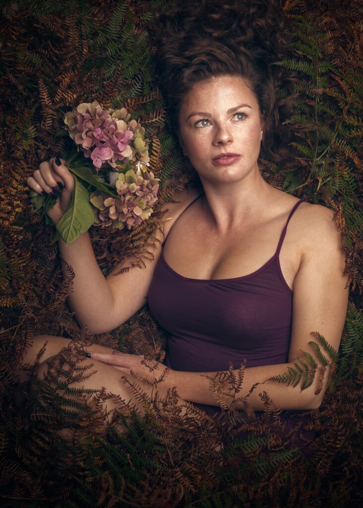 A woman in a burgundy top holds pink hydrangeas, surrounded by ferns in autumnal tones, looking directly at the camera.