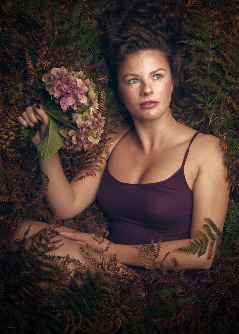 A woman in a burgundy top holds pink hydrangeas, surrounded by ferns in autumnal tones, looking directly at the camera.
