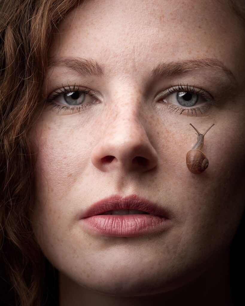 A woman's face in close-up with a snail crawling on her cheek. Her expression is neutral and contemplative.