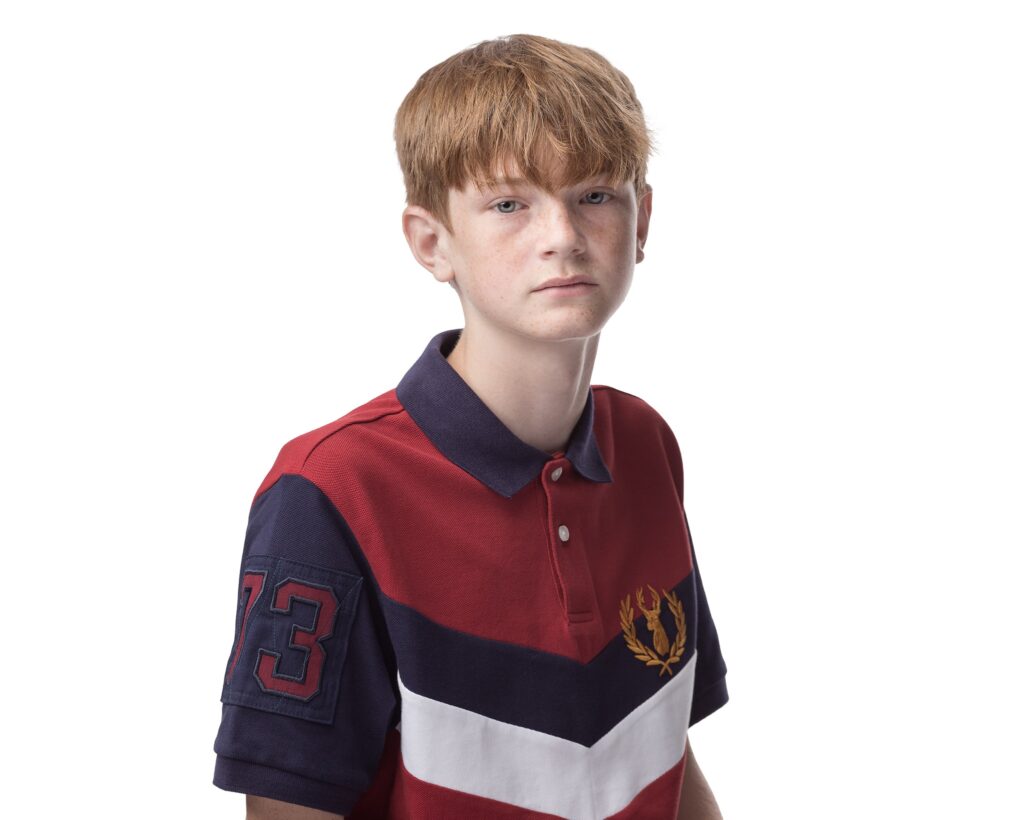 A young boy with reddish-brown hair wearing a red and navy polo shirt with a number 13 patch, against a white background.