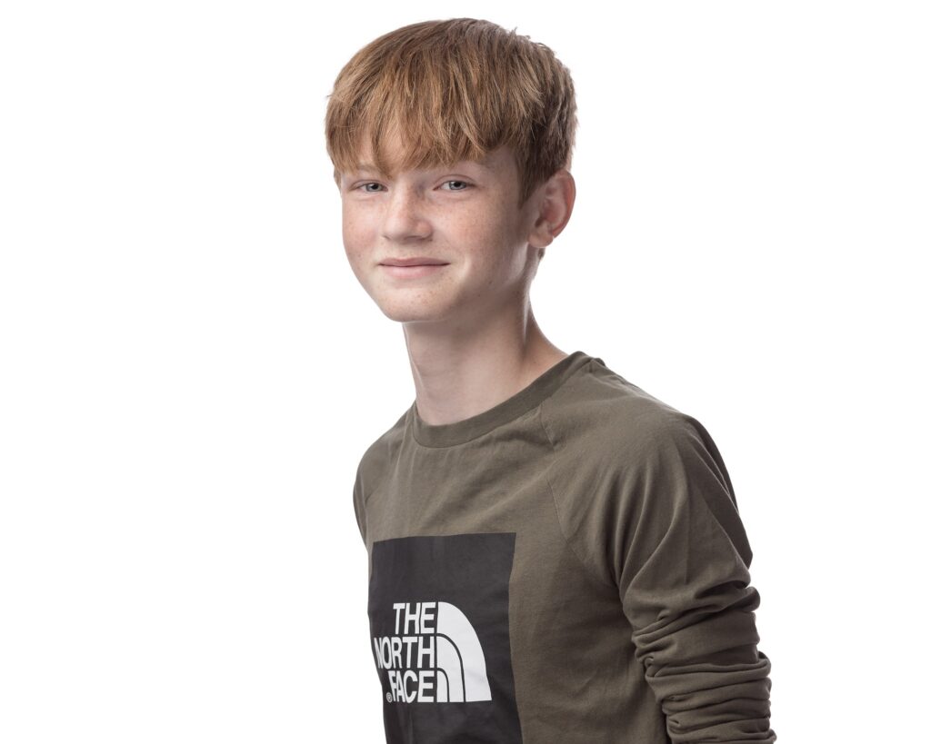 A young boy with reddish-brown hair wearing a khaki and black The North Face t-shirt, smiling at the camera against a white background.