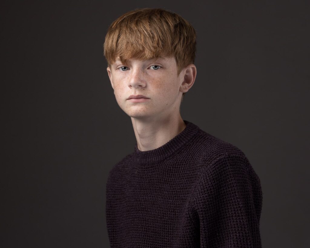 A young person with reddish-brown hair and freckles, wearing a dark knit jumper, looks directly at the camera against a grey background.